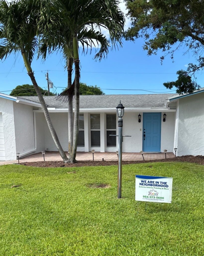 Front of a white house with a bright blue door, palm trees, and a yard sign in the lawn reading 'We Are in the Neighborhood' with a contact number and logo.