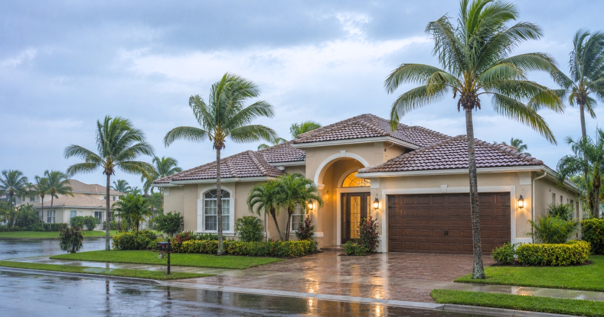 Beige single-story house with a brown tile roof, arched entry, and two-car garage, surrounded by palm trees on a wet street in a tropical neighborhood.