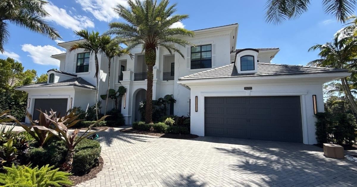White two-story home with a gray-tiled roof, arched entry, and a two-car garage surrounded by palm trees on a sunny day.