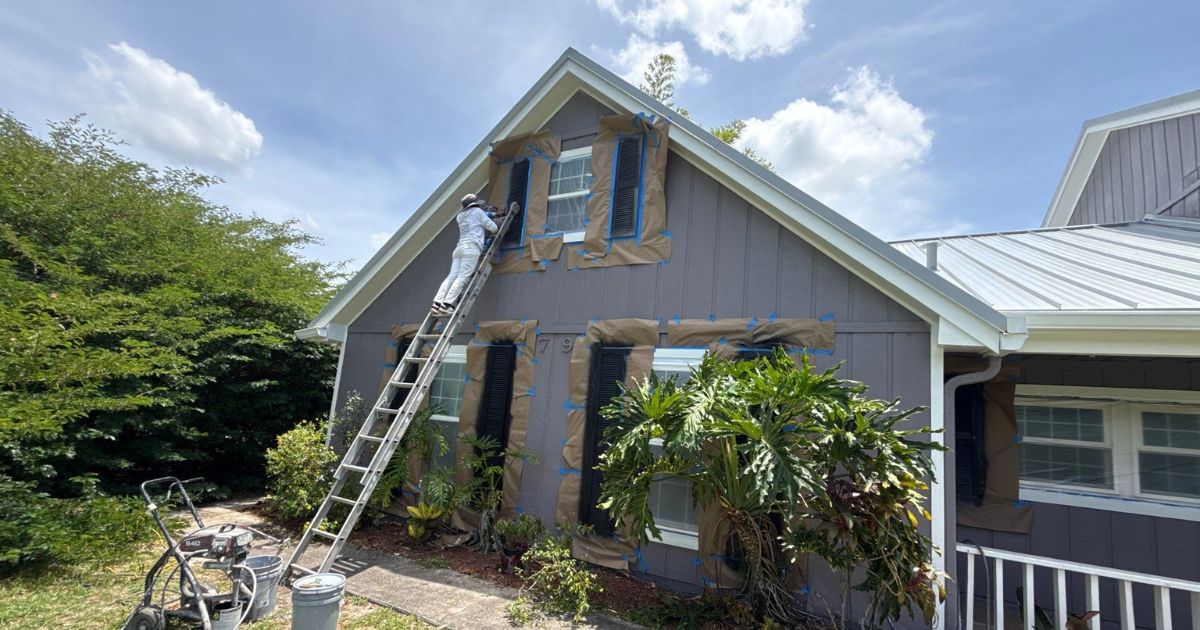 Person on a ladder taping and papering windows on a gray suburban house for exterior painting, with tools on the yard.