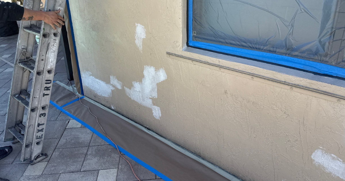 Exterior Wall prep in florida - Tru Colors Contracting Ladder against a beige stucco wall while white patch filler dries; window masked with plastic and blue tape, drop cloth along the base.