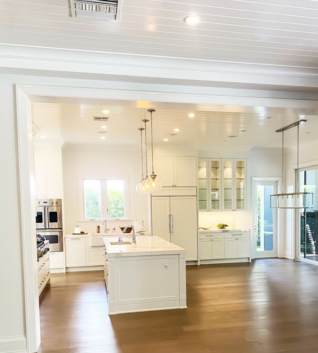 Bright white kitchen with an island, pendant lights, and glass-front cabinets; warm hardwood floor and large windows bringing in natural light