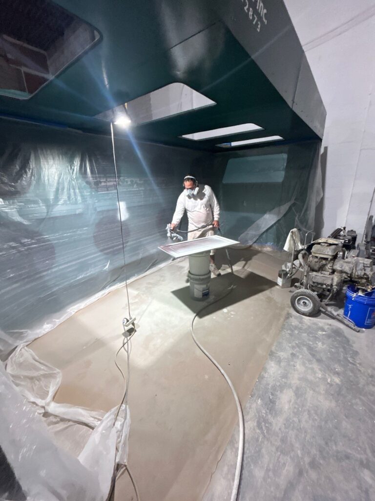 Worker in white protective suit and respirator spraying paint inside a plastic-walled booth; large teal structure overhead and hoses on the floor.
